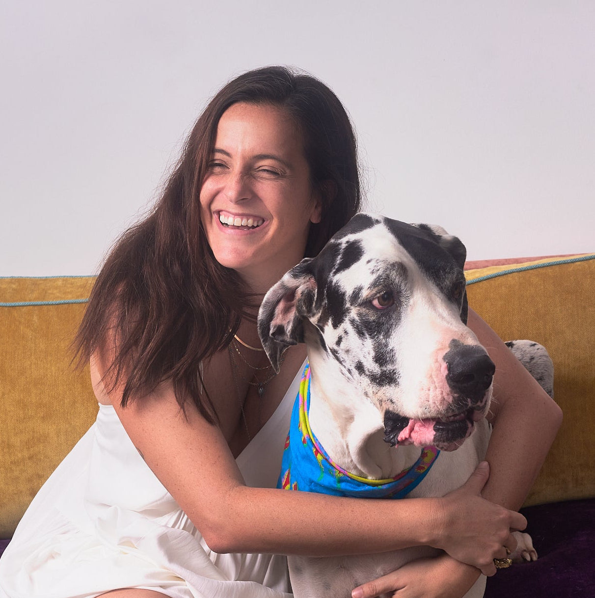 Woman in a white dress sitting on the floor with a large spotted dog on a yellow couch.