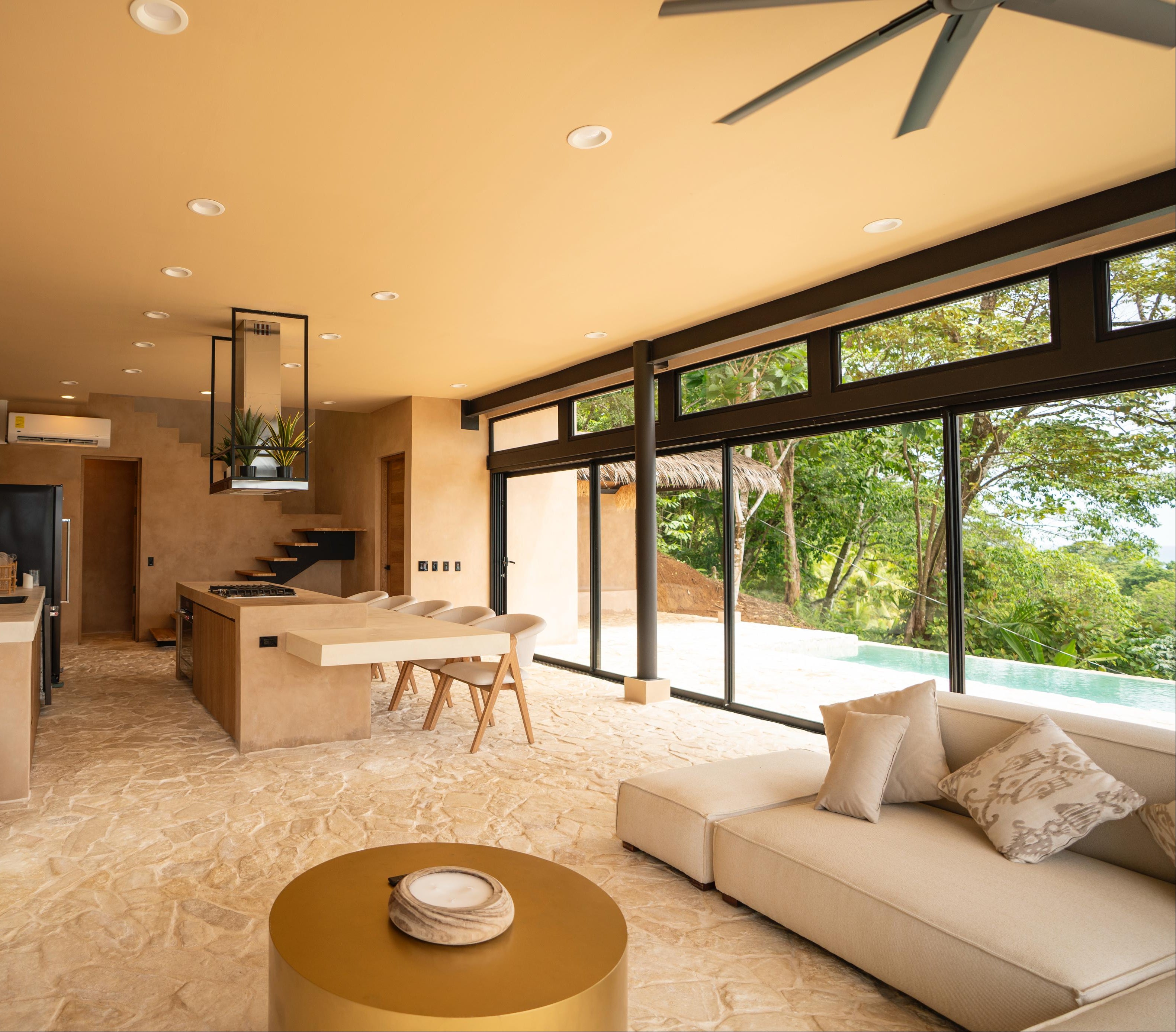 Modern living room with a kitchen in the background, featuring a couch, coffee table, and large windows.
